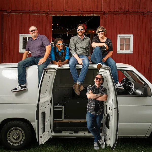 A group of five people posing with a white van containing audio equipment in front of a red barn venue.
