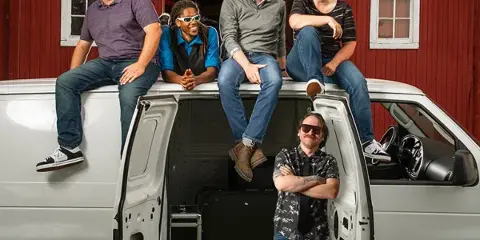 A group of five people posing with a white van containing audio equipment in front of a red barn venue.