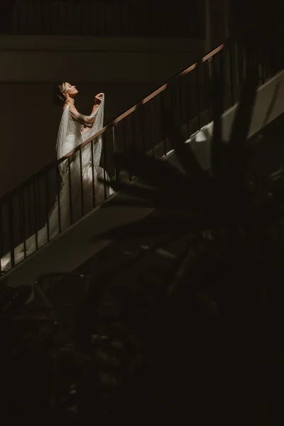 Bride in a white wedding gown standing on a staircase with dramatic lighting and palm fronds in the foreground.