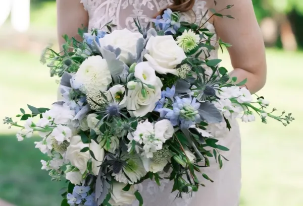 Bride in white lace gown holding a cascading wedding bouquet of white roses, blue delphiniums, and greenery outdoors.