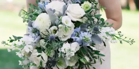Bride in white lace gown holding a cascading wedding bouquet of white roses, blue delphiniums, and greenery outdoors.