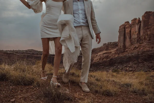 A bride and groom in white wedding attire standing in a red rock desert landscape.