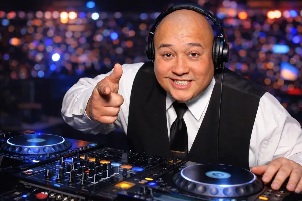 A smiling DJ wearing headphones and a vest stands behind a mixing console and turntables, pointing at the camera with a blurred crowd in the background.