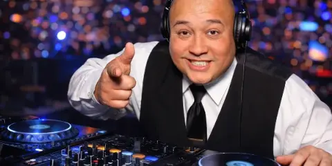 A smiling DJ wearing headphones and a vest stands behind a mixing console and turntables, pointing at the camera with a blurred crowd in the background.