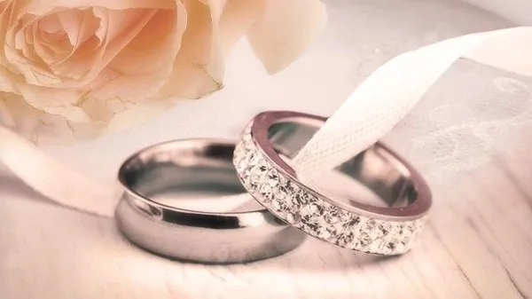 A pair of wedding rings, one plain and one with diamonds, resting on a white ribbon next to a pale rose.