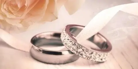 A pair of wedding rings, one plain and one with diamonds, resting on a white ribbon next to a pale rose.
