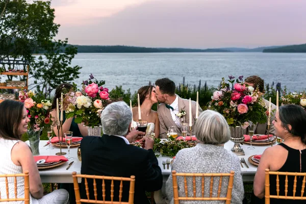 A wedding couple and guests seated at a reception table overlooking a coastal body of water at dusk.