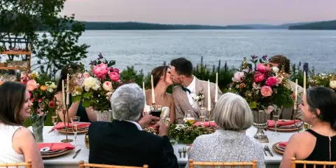 A wedding couple and guests seated at a reception table overlooking a coastal body of water at dusk.