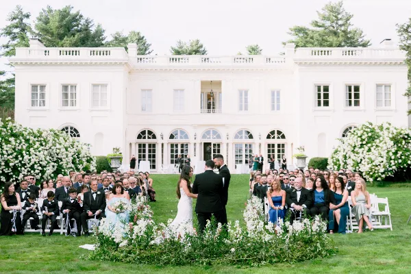 A wedding ceremony taking place on the expansive lawn in front of the white Wadsworth Mansion, with guests seated in white chairs and floral arrangements.