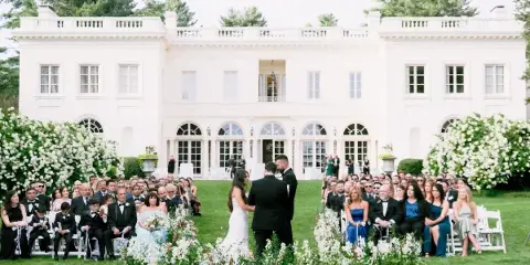 A wedding ceremony taking place on the expansive lawn in front of the white Wadsworth Mansion, with guests seated in white chairs and floral arrangements.