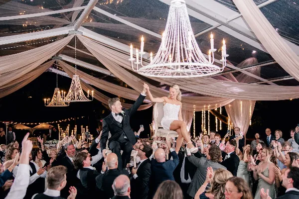A bride and groom dancing on a raised platform during a wedding reception, surrounded by a crowd of guests and illuminated by chandeliers.
