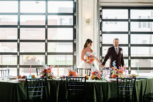 A bride and groom standing behind a decorated wedding reception table inside a venue with large industrial-style windows.