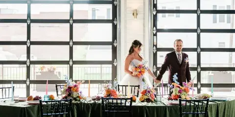 A bride and groom standing behind a decorated wedding reception table inside a venue with large industrial-style windows.