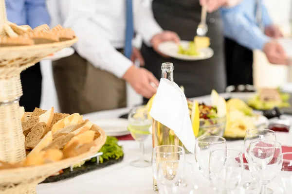 A catering buffet table with bread, wine, and food being served by waiters in a formal event setting.