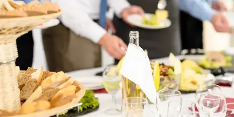 A catering buffet table with bread, wine, and food being served by waiters in a formal event setting.