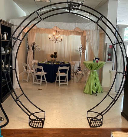 A black metal wedding arch set up in a showroom with a decorated table and chairs in the background.