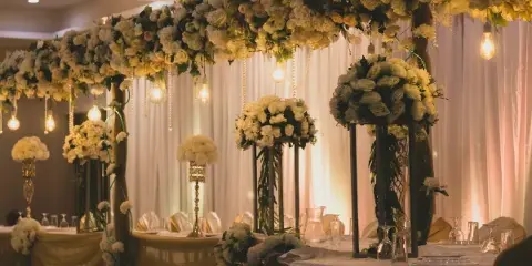 A decorated wedding reception table featuring white floral centerpieces, hanging lights, and draped fabric.