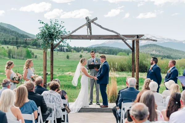 A bride and groom exchanging vows under a rustic wooden arch during an outdoor wedding ceremony in a green field with mountains in the background.