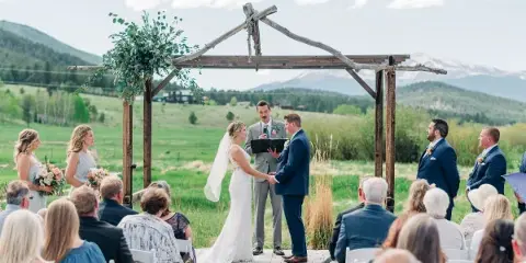 A bride and groom exchanging vows under a rustic wooden arch during an outdoor wedding ceremony in a green field with mountains in the background.