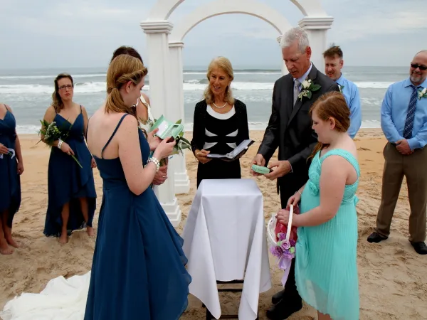 A wedding officiant presides over a beach ceremony where a father presents a medallion to a young girl, with the bride and groom standing nearby.