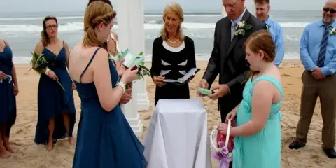 A wedding officiant presides over a beach ceremony where a father presents a medallion to a young girl, with the bride and groom standing nearby.