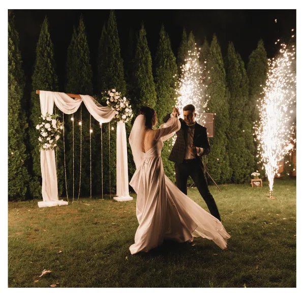A bride and groom dancing at night in front of a floral wedding arch with sparklers.