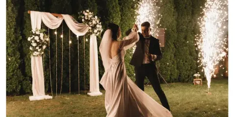 A bride and groom dancing at night in front of a floral wedding arch with sparklers.