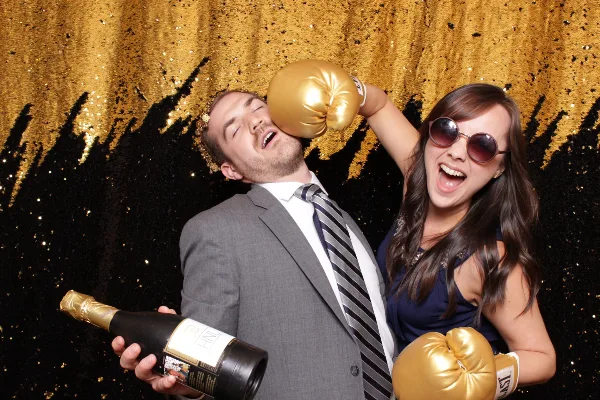 A man in a suit and a woman in a dress pose playfully with gold boxing gloves and a champagne bottle in front of a black and gold sequined photo booth backdrop.