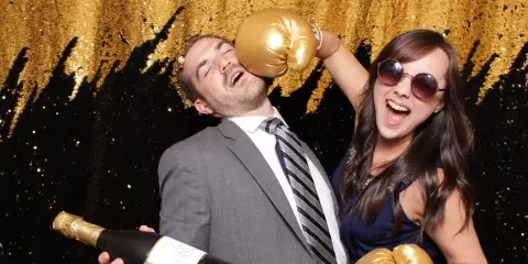 A man in a suit and a woman in a dress pose playfully with gold boxing gloves and a champagne bottle in front of a black and gold sequined photo booth backdrop.