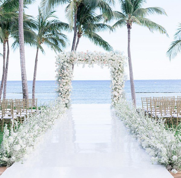 A white floral wedding arch and aisle set up on a beach with palm trees and the ocean in the background.