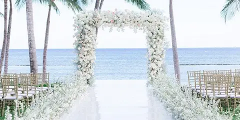 A white floral wedding arch and aisle set up on a beach with palm trees and the ocean in the background.