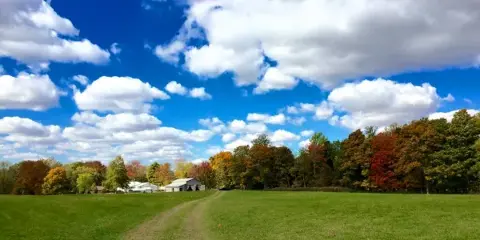 A wide shot of a rural property featuring a large green field, a dirt path, and a barn in the distance under a blue sky with clouds.
