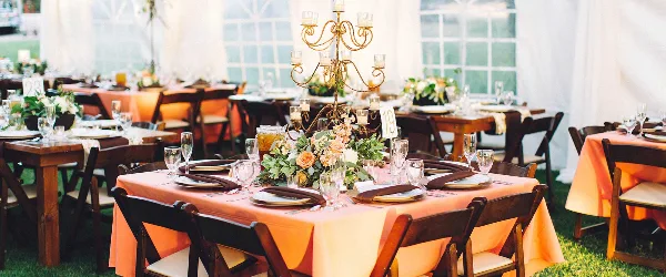 An outdoor event setup featuring wooden tables with orange linens, chairs, and floral centerpieces.