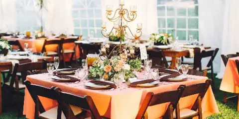 An outdoor event setup featuring wooden tables with orange linens, chairs, and floral centerpieces.