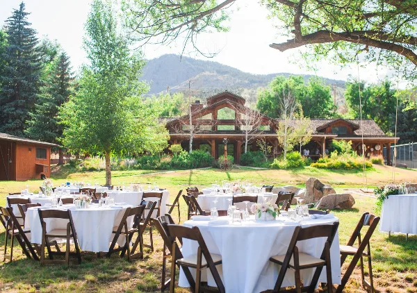 Outdoor wedding reception setup with white tables and chairs on a lawn in front of a rustic timber-frame building with mountains in the background.