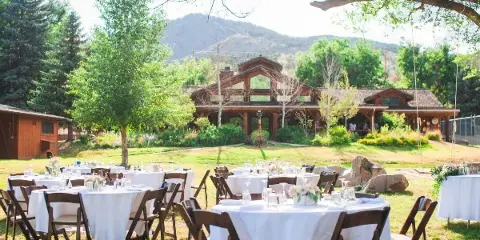 Outdoor wedding reception setup with white tables and chairs on a lawn in front of a rustic timber-frame building with mountains in the background.