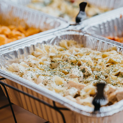Catering buffet with aluminum chafing dishes holding pasta with chicken and herbs, served with tongs.