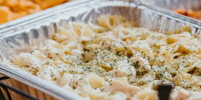 Catering buffet with aluminum chafing dishes holding pasta with chicken and herbs, served with tongs.