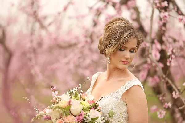 Bride with styled hair and makeup holding a floral bouquet in front of blooming trees