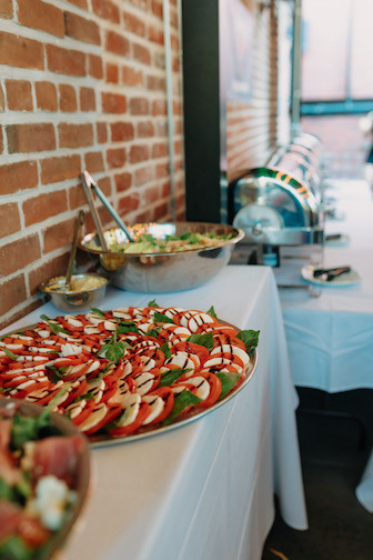 A catering buffet setup featuring a large platter of sliced fruit and vegetables, a bowl of salad, and chafing dishes on a white tablecloth.
