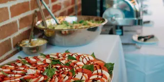 A catering buffet setup featuring a large platter of sliced fruit and vegetables, a bowl of salad, and chafing dishes on a white tablecloth.