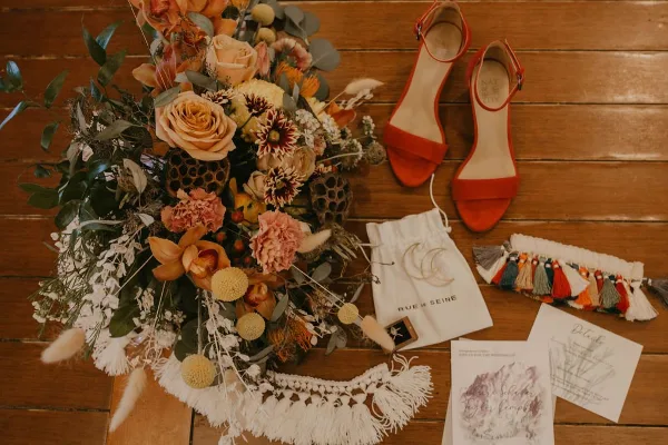 A detailed overhead shot of a wedding bouquet with orange roses and dahlias, placed on a wooden table next to red heels and wedding stationery.