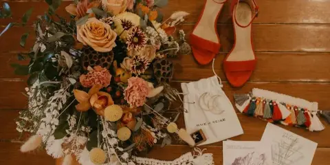 A detailed overhead shot of a wedding bouquet with orange roses and dahlias, placed on a wooden table next to red heels and wedding stationery.