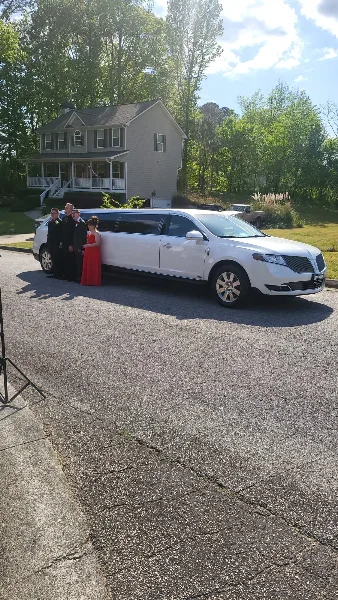 A white stretch limousine parked on a driveway with a group of formally dressed people standing beside it in front of a house.