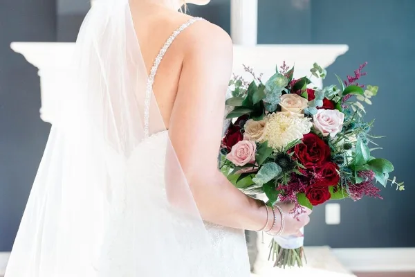 A bride in a white lace dress and veil holding a large, mixed floral bouquet with red and cream roses.