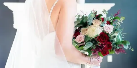 A bride in a white lace dress and veil holding a large, mixed floral bouquet with red and cream roses.