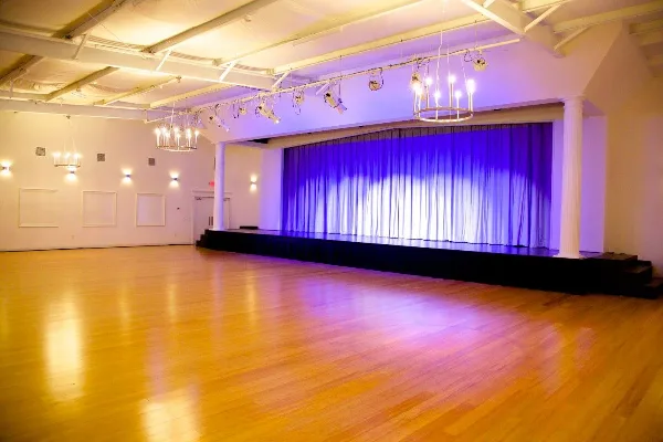 Interior view of a large event venue featuring a wooden dance floor, a stage with blue curtains, and hanging chandeliers.