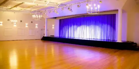 Interior view of a large event venue featuring a wooden dance floor, a stage with blue curtains, and hanging chandeliers.
