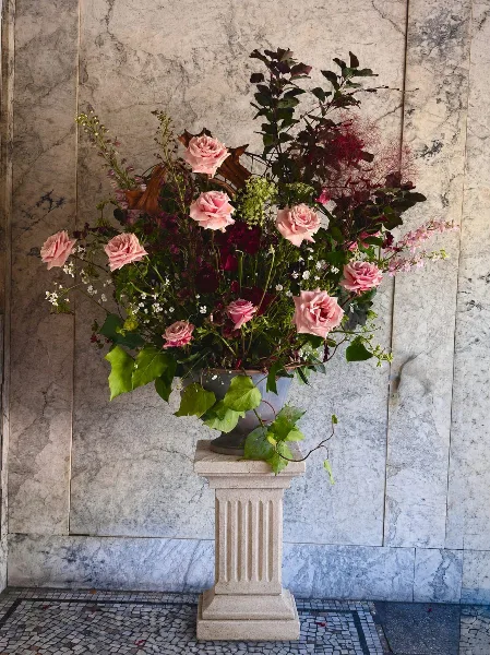 A large floral arrangement with pink roses and greenery in a ceramic vase on a stone pedestal against a marble wall.