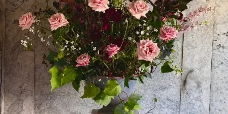 A large floral arrangement with pink roses and greenery in a ceramic vase on a stone pedestal against a marble wall.